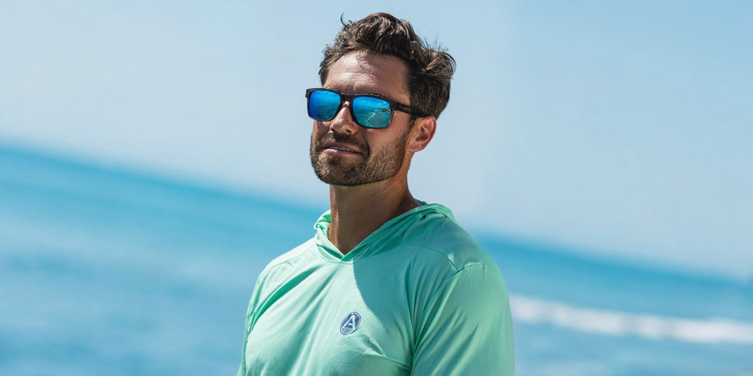 A man wearing a mint green hooded long-sleeve athletic shirt and blue mirrored sunglasses, standing outdoors with a blurred ocean and blue sky background.